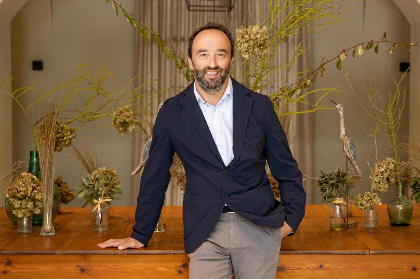Smiling man with beard wearing a navy blazer and light blue shirt, leaning on a wooden table with dried floral arrangements and decorative birds behind him.