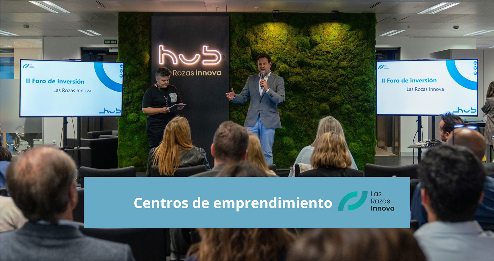 Man in a gray blazer speaking into a microphone at Las Rozas Innova investment forum with audience seated and two screens displaying event title.