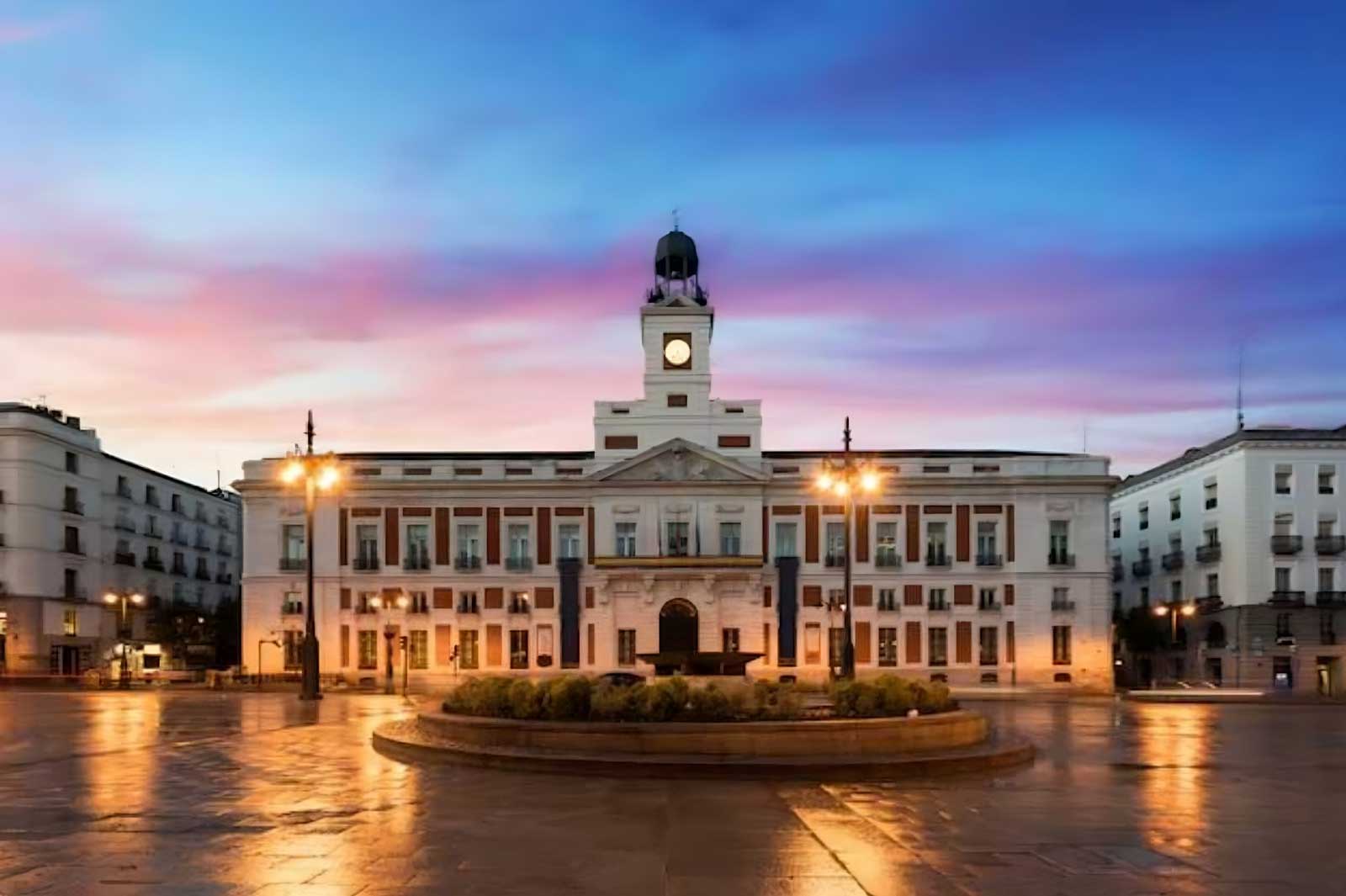 Historic white clock tower building at sunset with illuminated street lamps reflecting on wet pavement in a public square.