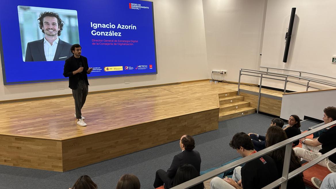 Man giving a presentation on stage with a large screen behind showing Ignacio Azorín González and his title in Spanish, while an audience seated in tiered seats watches.