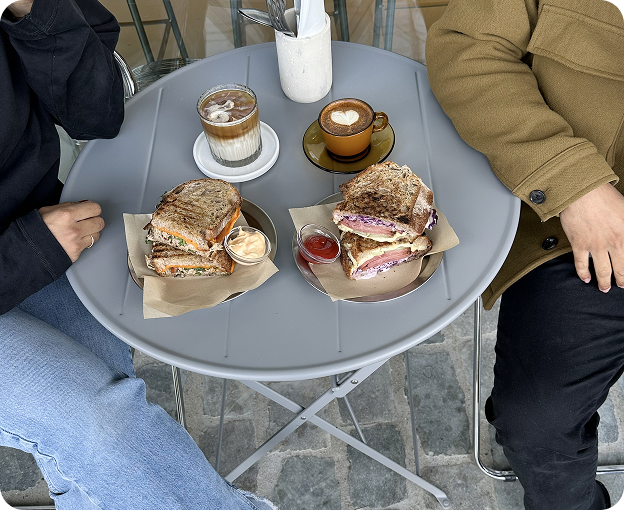 Small outdoor table with coffee, matcha and toasted sandwiches, shared by two people.
