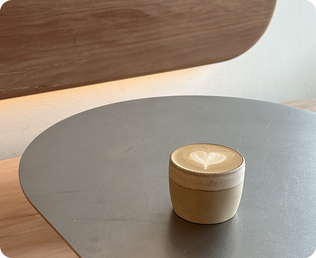 A ceramic cup of flat white with heart-shaped latte art, placed on a round grey table with warm wood tones in the background.