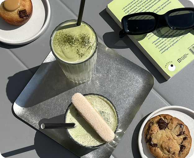 Matcha drinks served on a silver tray with a ladyfinger, surrounded by a cookie, muffin, sunglasses and a green book on a grey table in natural sunlight.