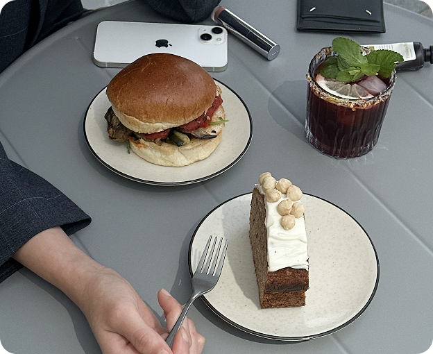 Lunch scene with a burger, slice of cake and iced drink on a grey table, with a hand and phone nearby.