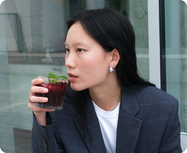 Woman drinking a dark iced cocktail outside Kosto, wearing a navy jacket.