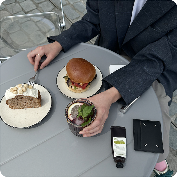 A lunch setup with a burger, salad bowl, and a cocktail on a grey table, paired with sunglasses and a phone.