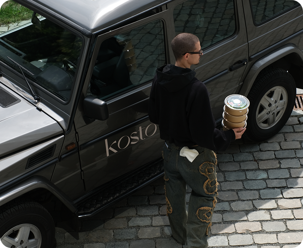 Man walking back to a Kosto-branded 4x4 with takeaway drinks in hand.