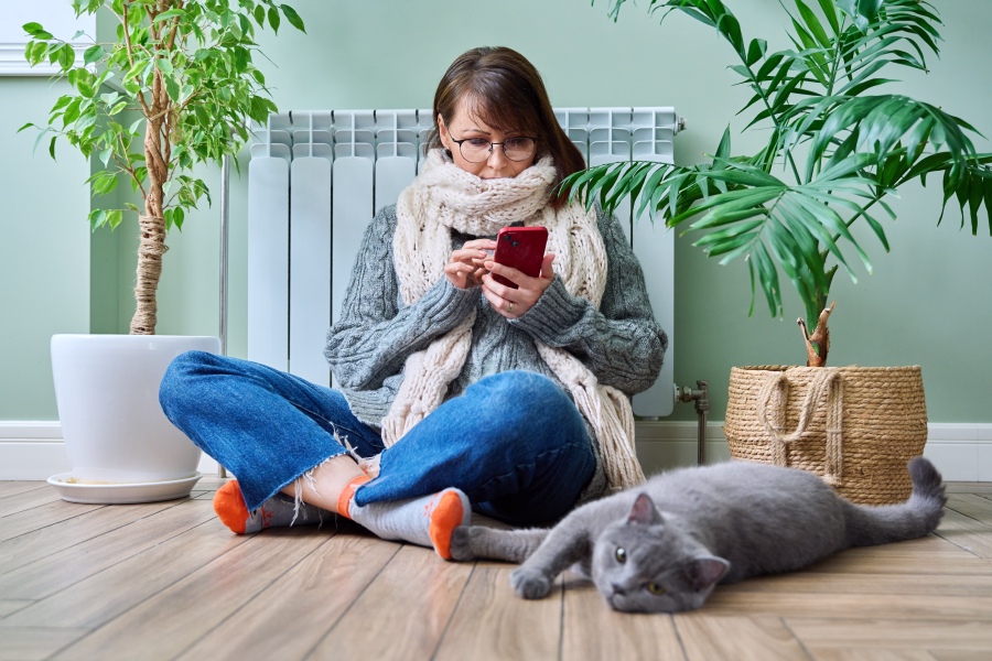 Person mit dickem Schal sitzt vor einem Heizkörper und bedient ein Smartphone.