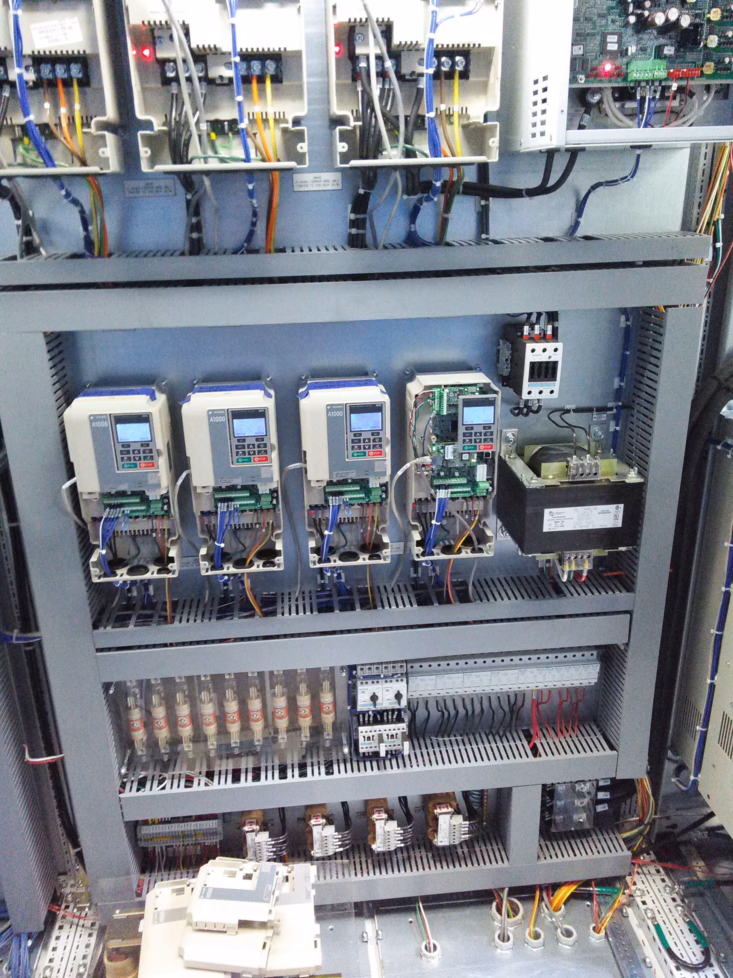 Electrical control panel with four A1000 variable frequency drives, circuit breakers, relays, wiring, and other electrical components mounted on a metal frame.