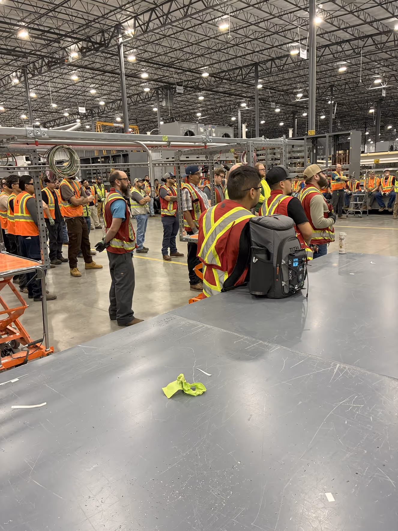 Group of workers wearing orange safety vests gathered inside a large industrial warehouse.