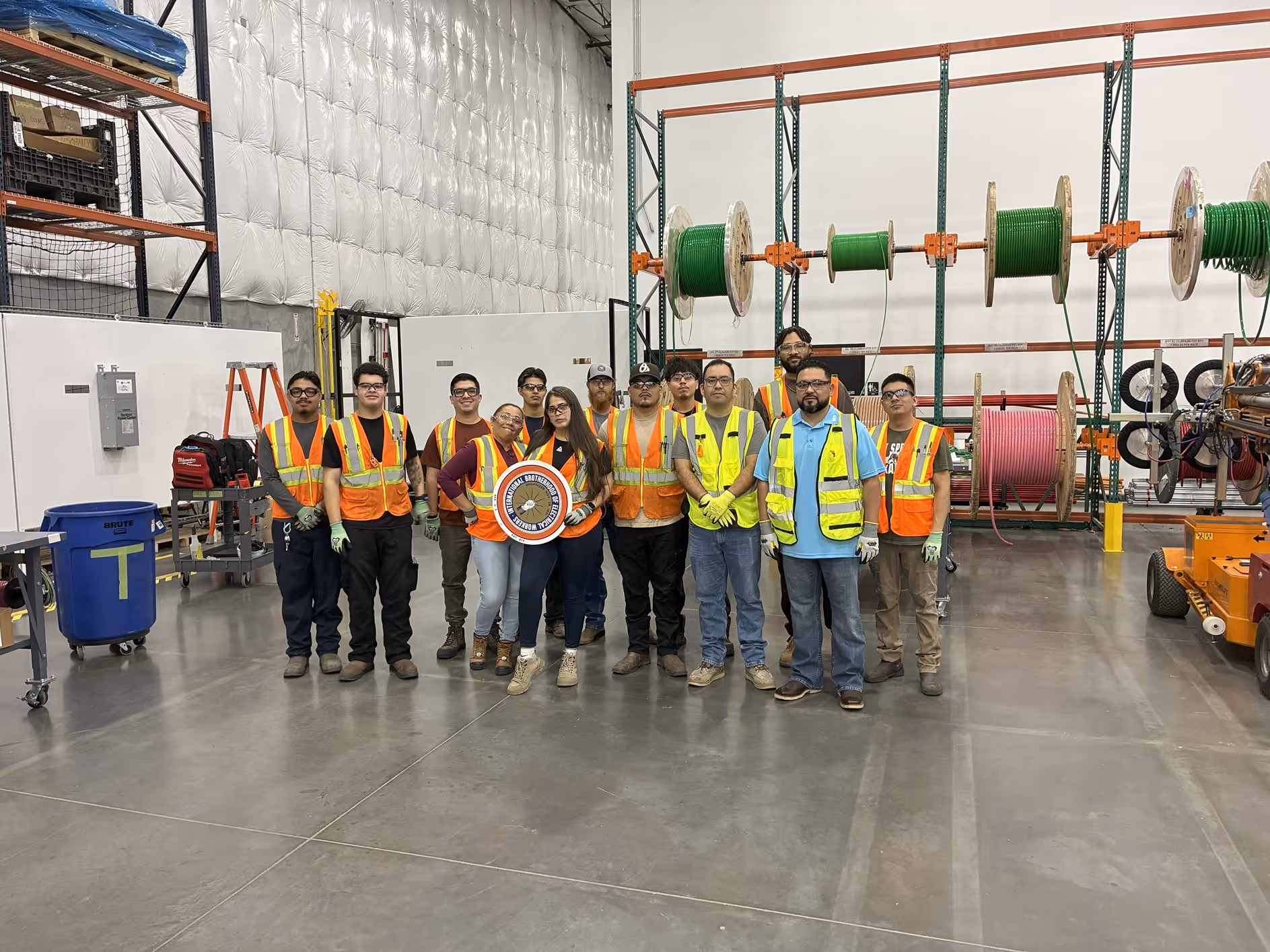 Group of workers wearing safety vests and gloves standing in a warehouse with industrial spools of cable behind them.