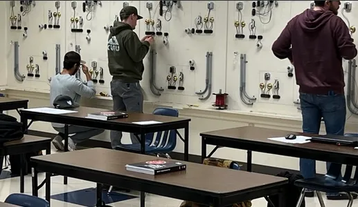 Three individuals working on electrical wiring setups on a training wall in a classroom with desks and chairs.