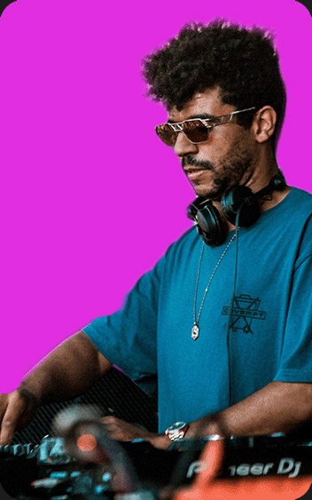 DJ Jamie Jones performing on a Pioneer DJ setup, wearing sunglasses and a blue t-shirt, with headphones around his neck, set against a vibrant pink background