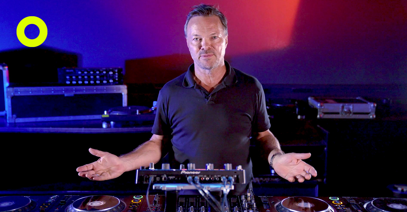 Pete Tong presenting a DJ lesson in a professional studio, standing behind a Pioneer DJ setup with a focused expression