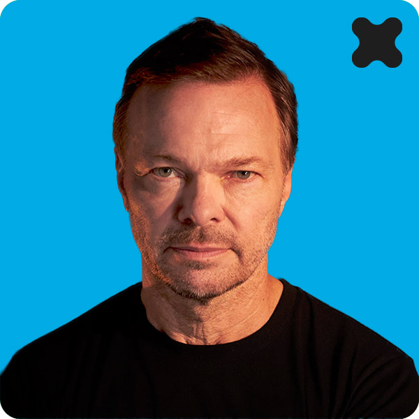 Portrait of DJ Pete Tong wearing a black shirt, set against a bright blue background