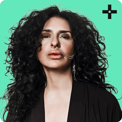 Portrait of DJ Nicole Moudaber with voluminous curly hair, wearing a black outfit and a statement nose chain, set against a mint green background