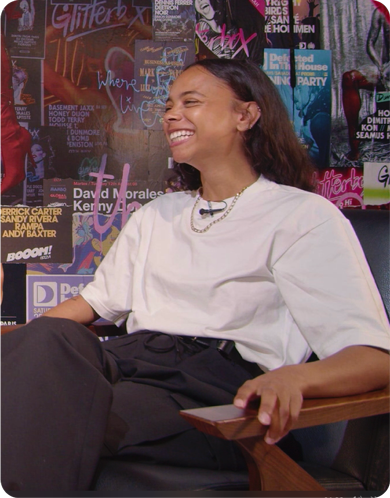 Smiling woman in a white t-shirt sitting in a chair during an interview, with a colorful music event poster wall in the background