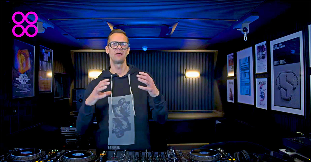 DJ instructor in a professional studio, standing behind a DJ setup and explaining a concept. The room features framed music posters and soundproofing panels