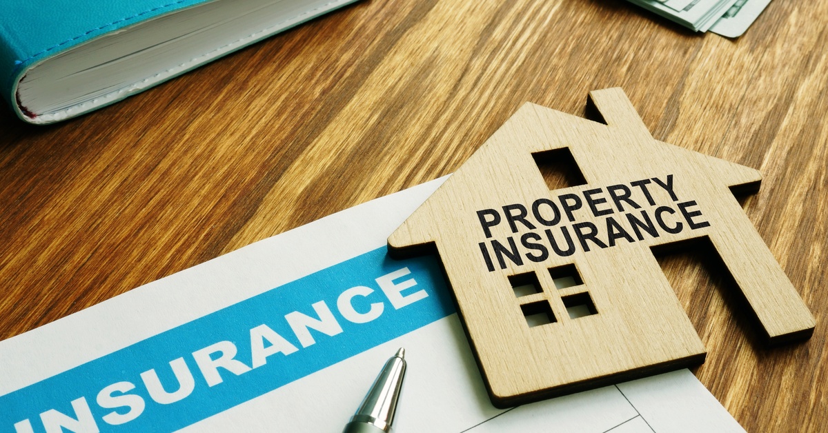 A document labeled INSURANCE sits on a wooden desk with cash, a pen, and a wooden house reading PROPERTY INSURANCE.