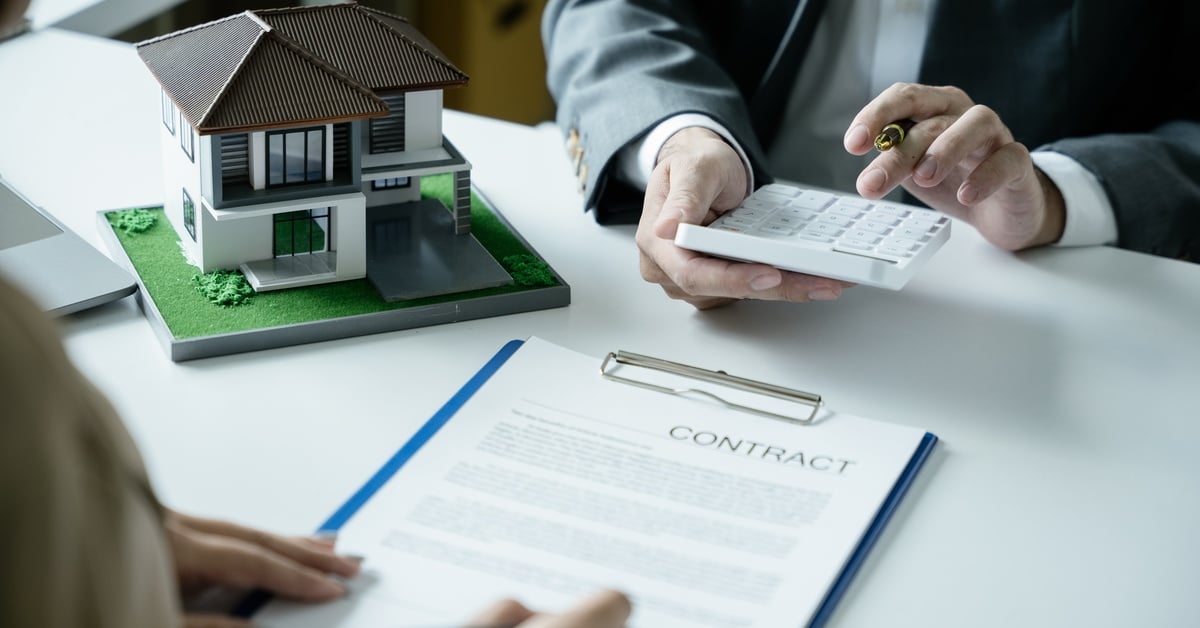 Two people sitting across a desk reviewing a home contract near a house model, calculator, and clipboard.