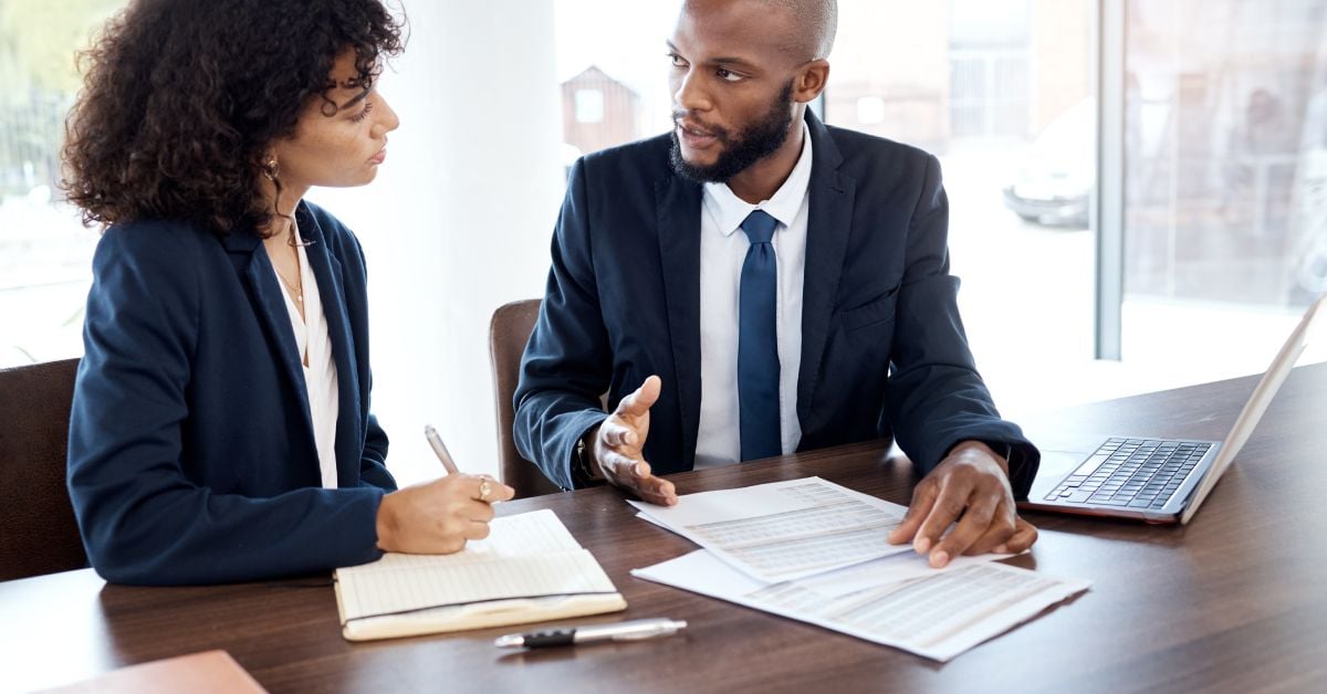 Two professionals sitting at a desk discussing financial documents while writing notes and using a laptop nearby.