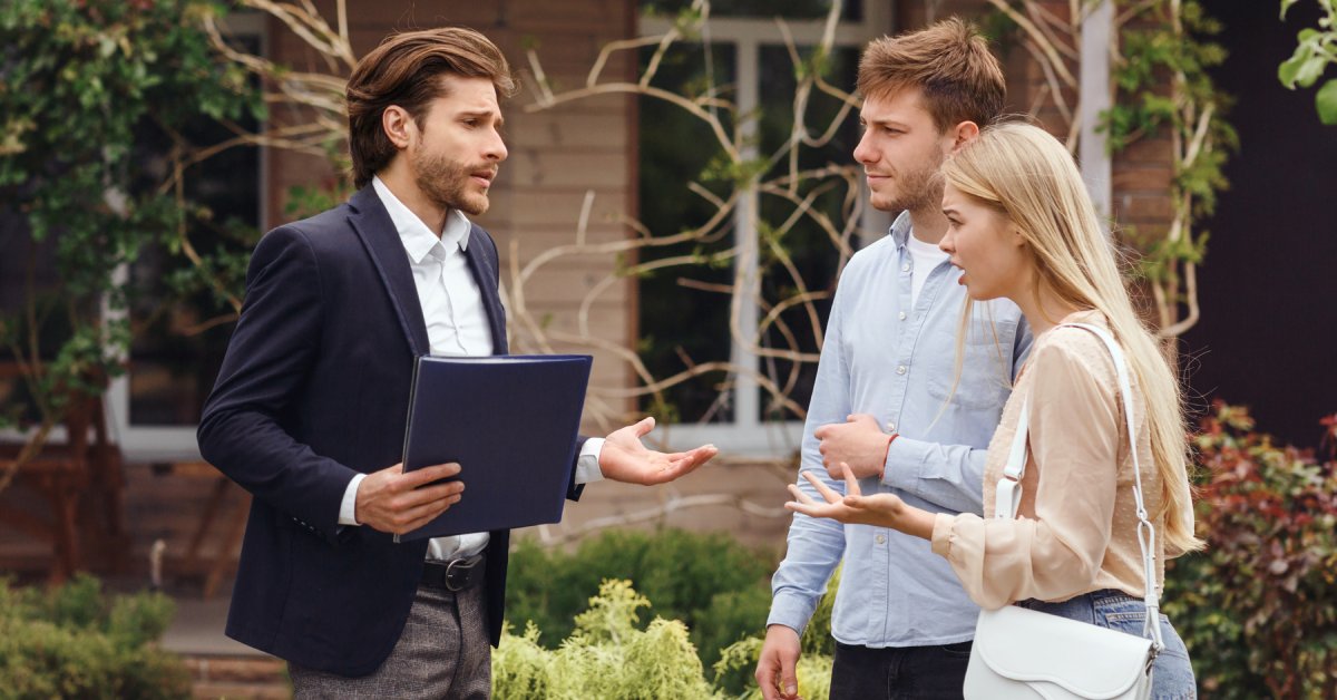 A man in a blazer holds a folder while talking to a young couple standing outside a residential building.