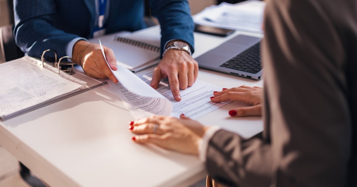 Two people at a desk with one person pointing at a document beside a laptop and a binder of paperwork.