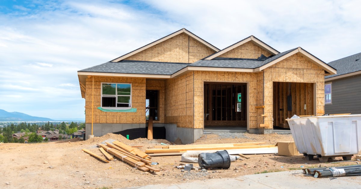 A house under construction with wood sheathing, roof shingles, a white dumpster, and scattered lumber in front.
