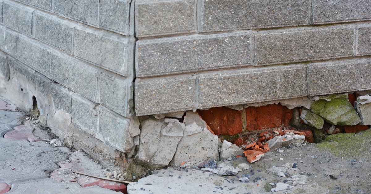Cracked concrete crumbling from a home’s corner foundation, revealing red brick and exposed mortar underneath.