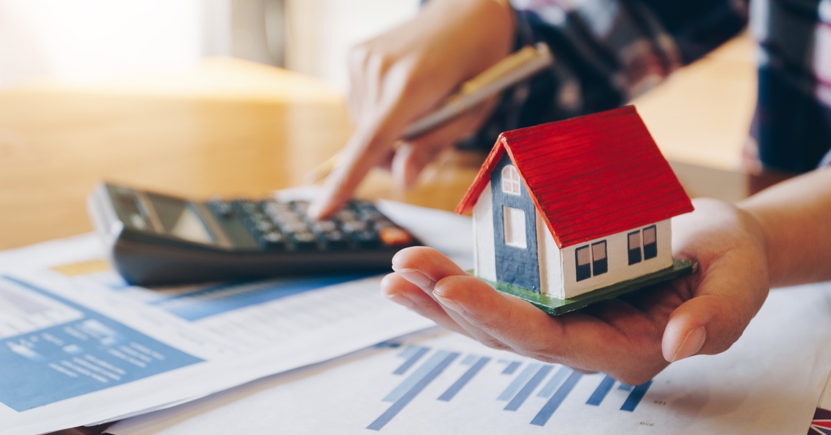 A person holding a small model house over financial documents as a calculator and pen rest nearby on the desk.