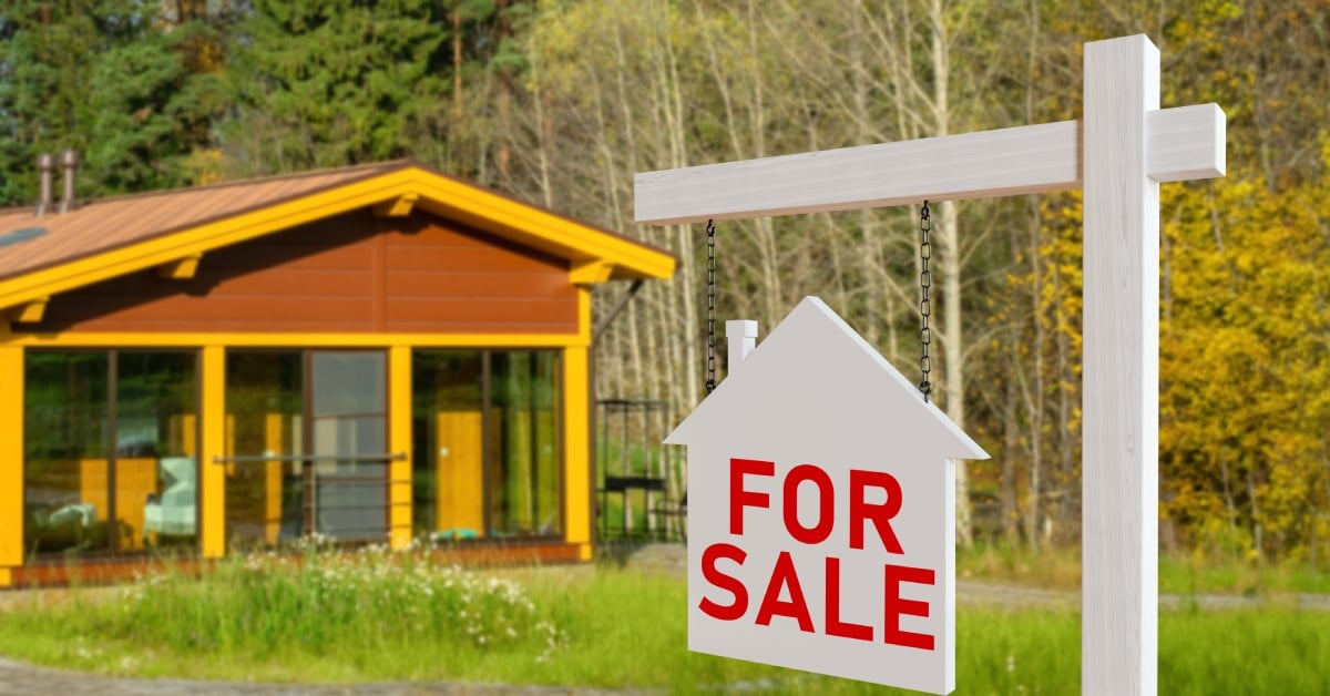 A house-shaped for sale sign hanging on a post in front of a wood cabin surrounded by trees in autumn color.