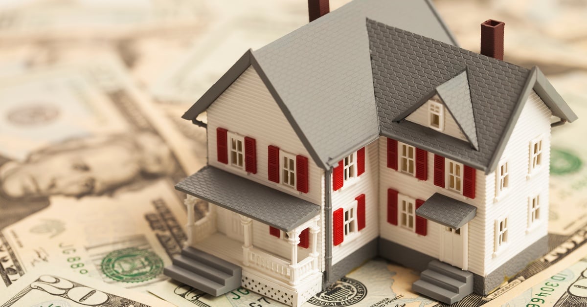 A plastic miniature of a home with a gray roof and red window shutters is resting over a pile of twenty dollar bills.