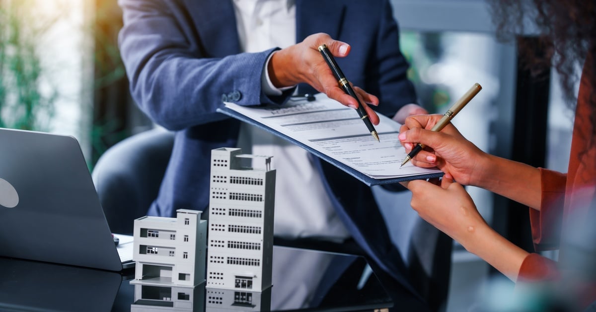 A woman signing paperwork on a clipboard. She is sitting across a desk from a man in a suit and there is a laptop on the desk.
