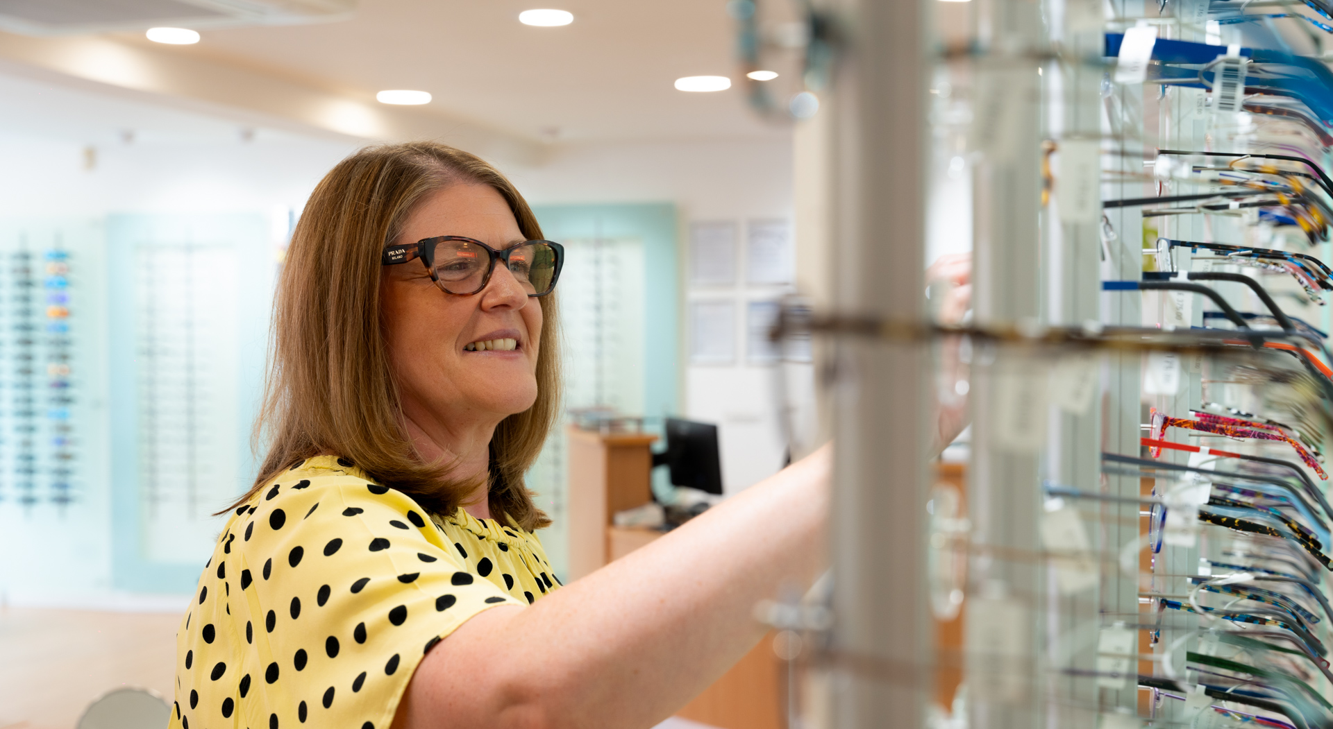 Woman in glasses smiling and selecting eyeglass frames in an eyewear store.
