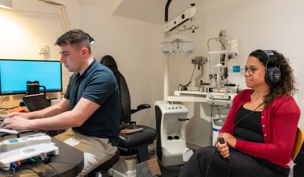 Audiologist operating computer while woman wearing headphones undergoes hearing test in clinic.