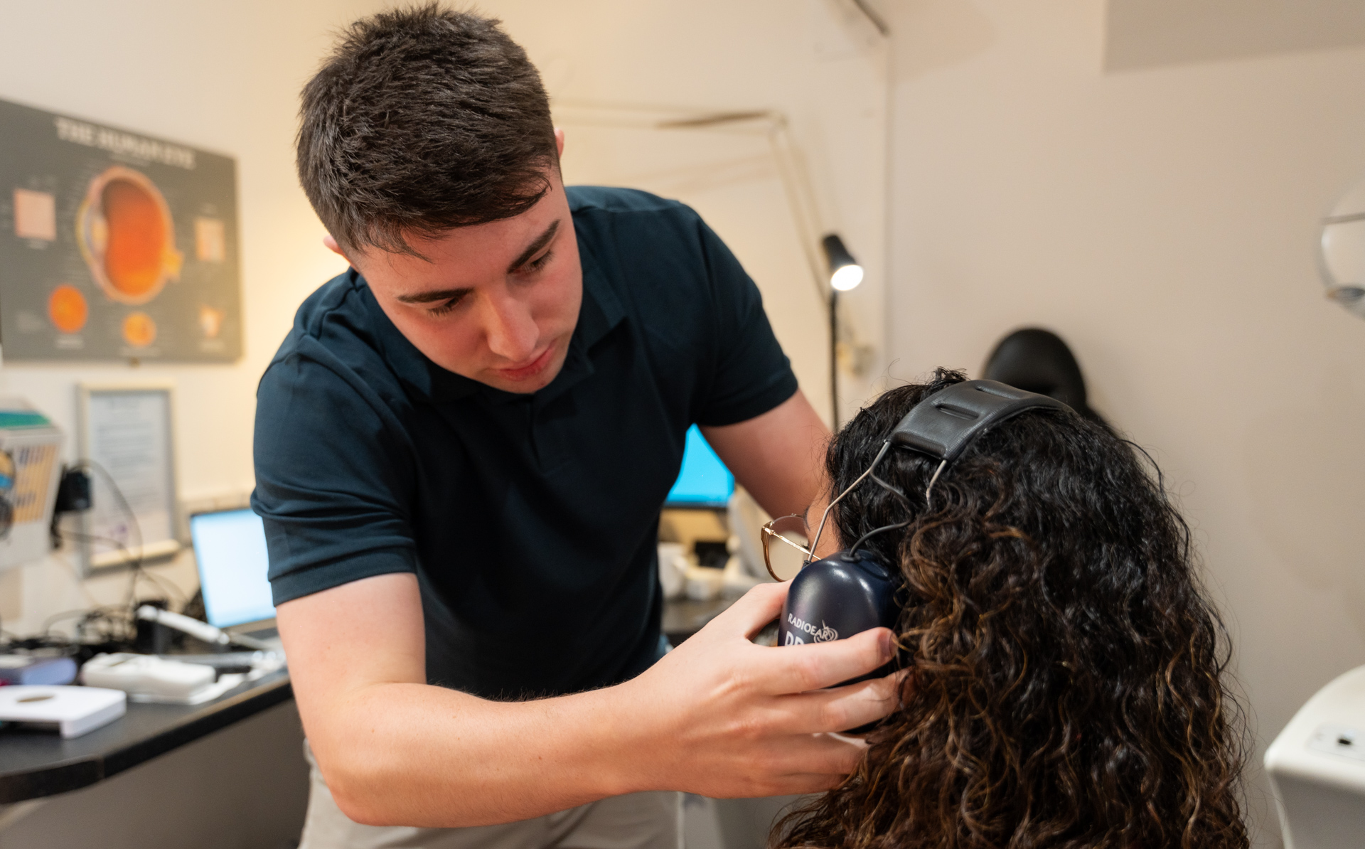 Audiologist fitting headphones on a seated woman with curly hair in a clinic room.