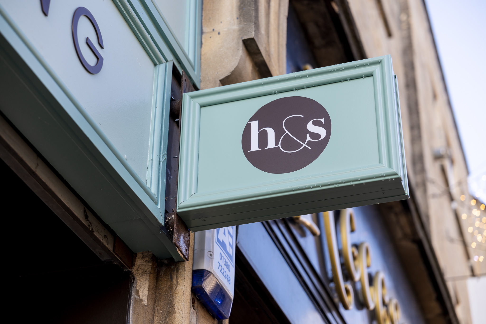 Rectangular mint-green sign with black circle logo and white letters h&s on building exterior.