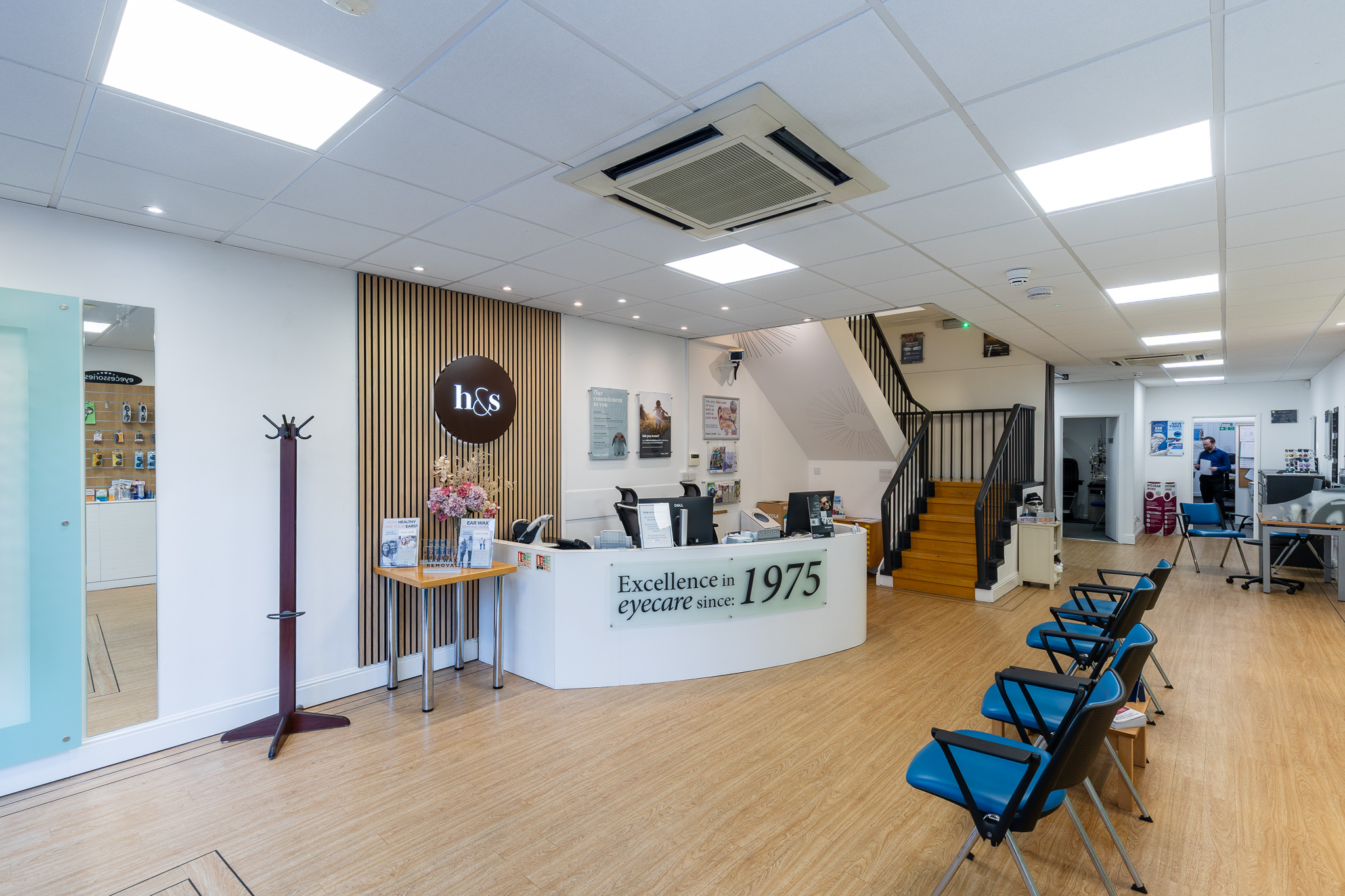 Reception area in an eyewear clinic with a white desk, seating, stairs, and a man in the background.