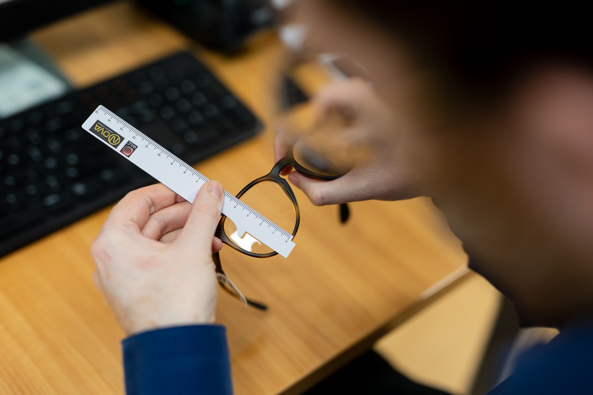 Person measuring eyeglass lens width with a white ruler over a wooden desk.
