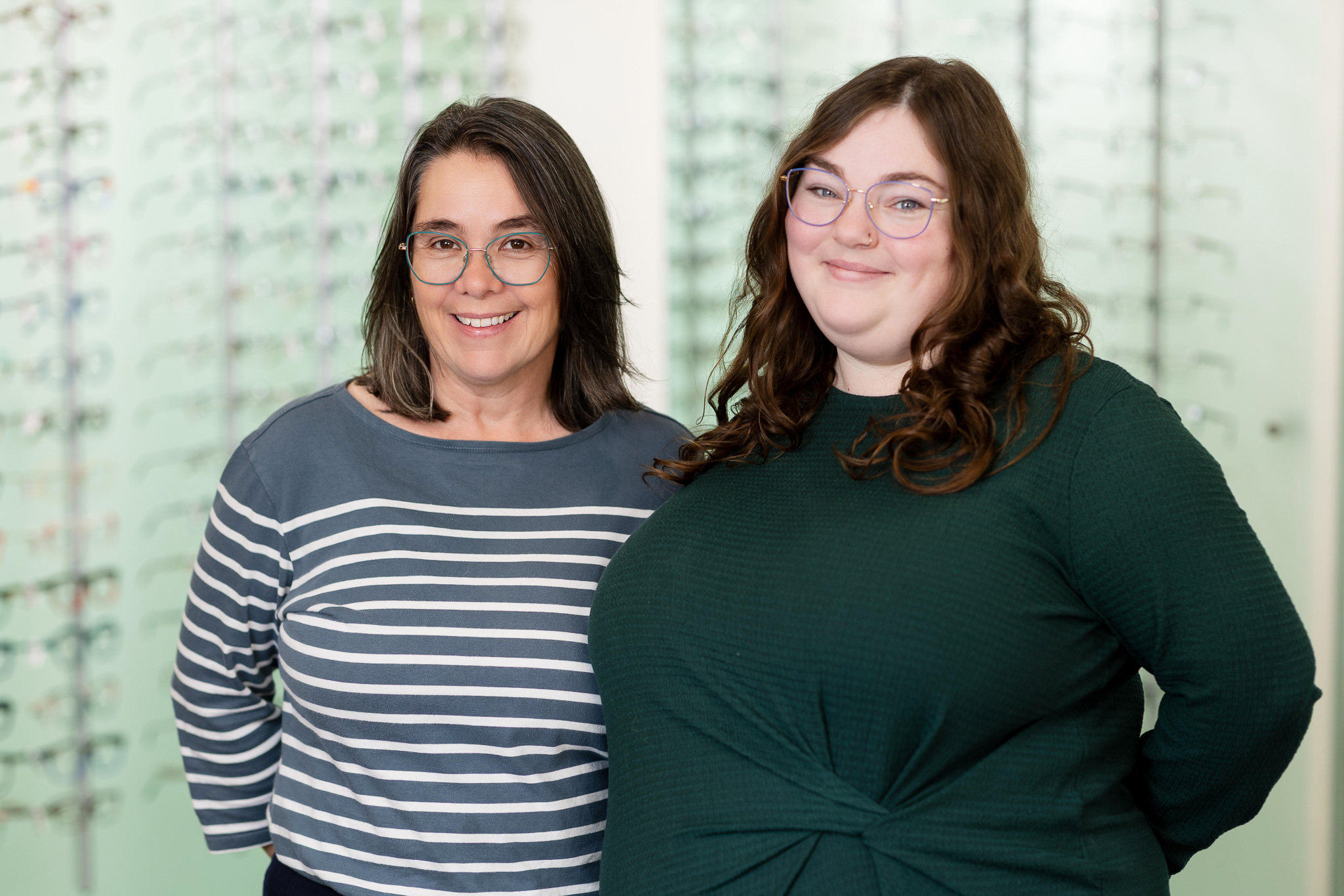 Two smiling women wearing glasses stand in an optician shop with eyeglass frames displayed behind them.