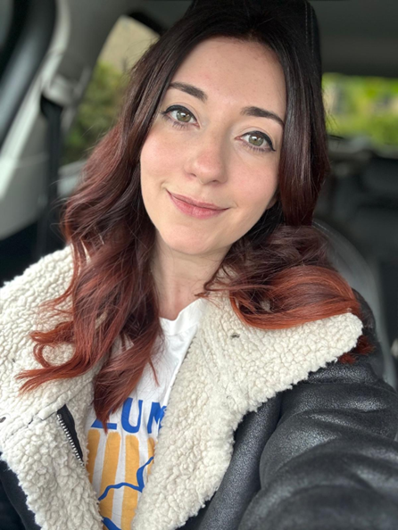 Woman with long auburn hair wearing a shearling jacket, smiling inside a car.