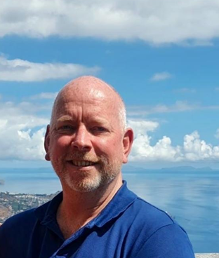 Smiling man with beard wearing a blue shirt, standing outdoors with sea and cloudy sky behind him.