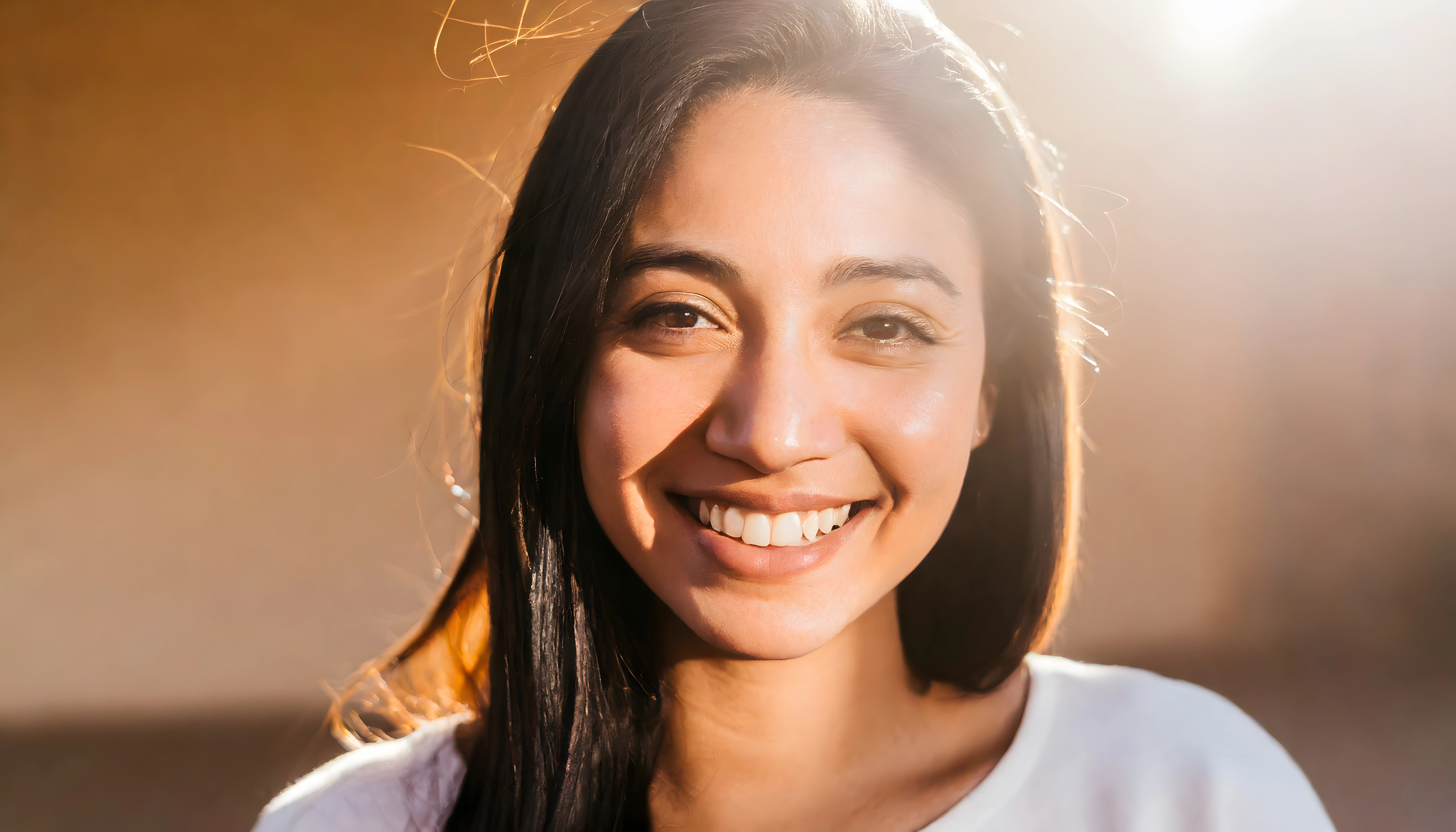 Smiling woman with long dark hair glowing in warm sunlight, wearing a white shirt.