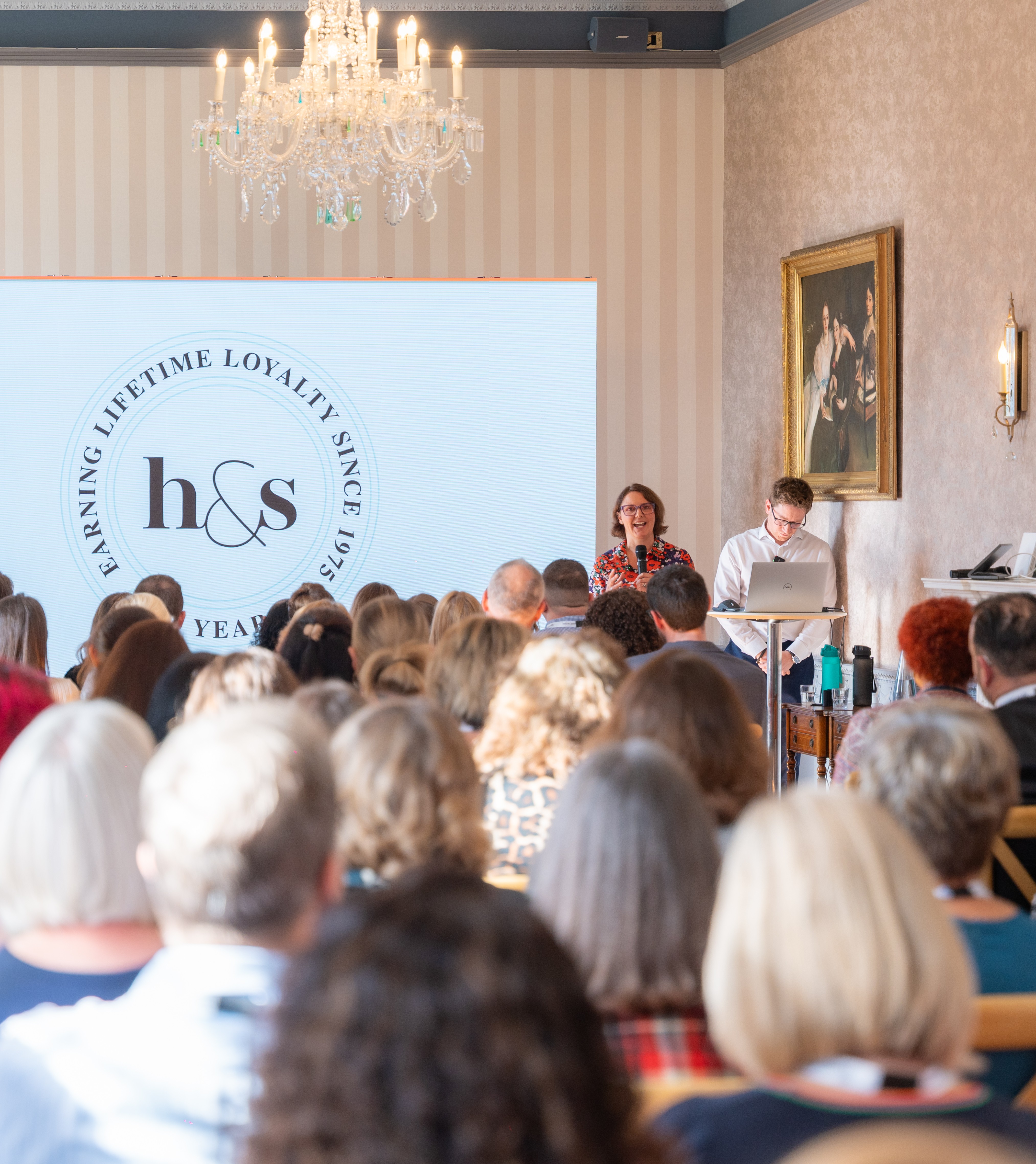 Woman presenting to a seated audience at an h&s 50th anniversary event, with a screen displaying the "Earning Lifetime Loyalty Since 1975" logo