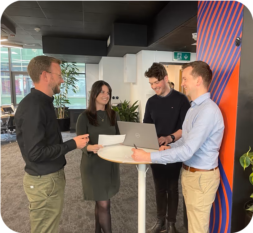 Four colleagues standing around a tall round table in a modern office, discussing papers and a laptop with smiles.