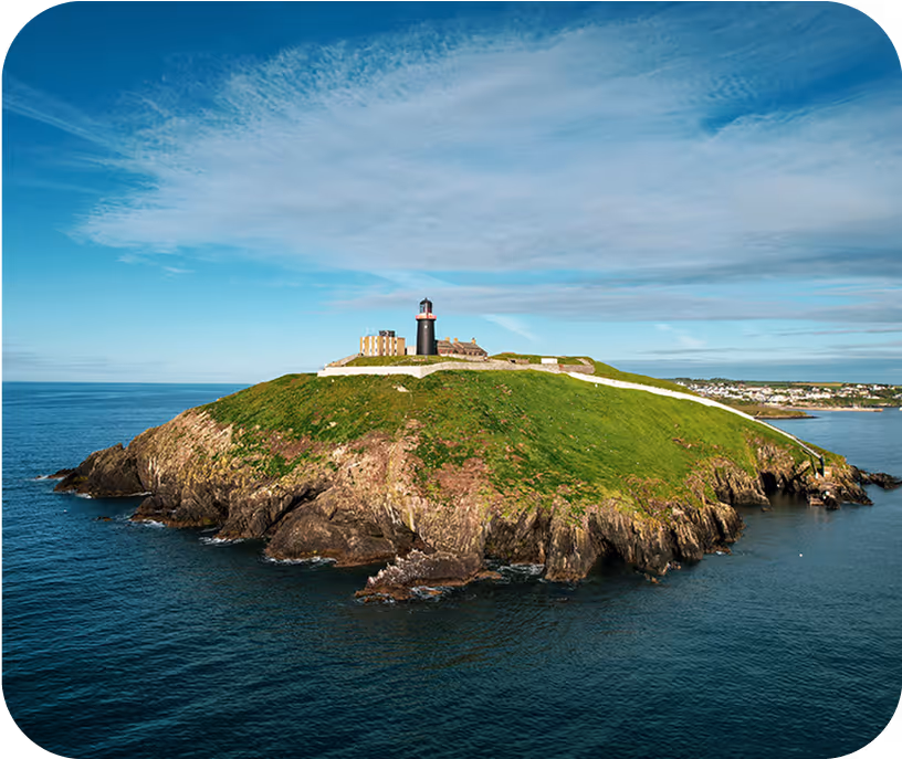 Small rocky island with green grass and a black lighthouse near a coastline under a partly cloudy blue sky.