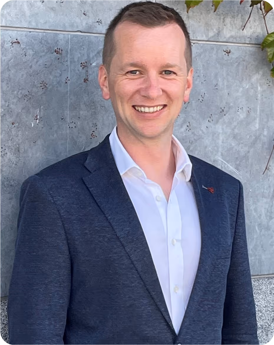 Smiling man wearing a dark blazer and white shirt standing against a textured gray wall with some green leaves visible in the top right corner.