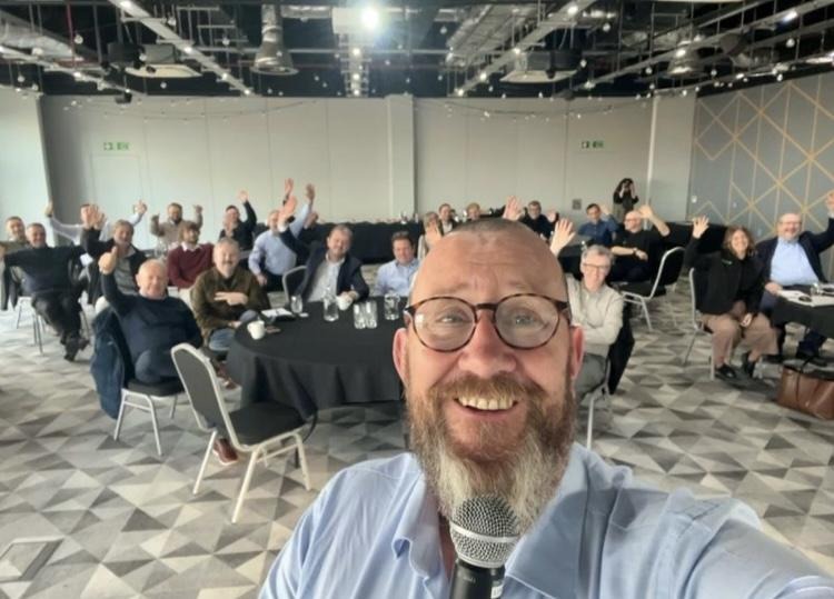 A person taking a selfie with a microphone in front of a group of peopleAI-generated content may be incorrect.