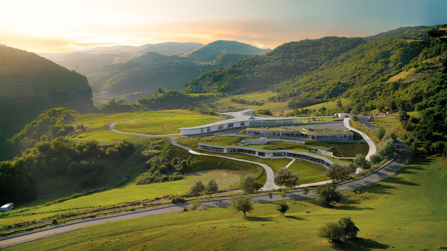 Modern building with curved white structures in a green valley surrounded by forested hills at sunset.