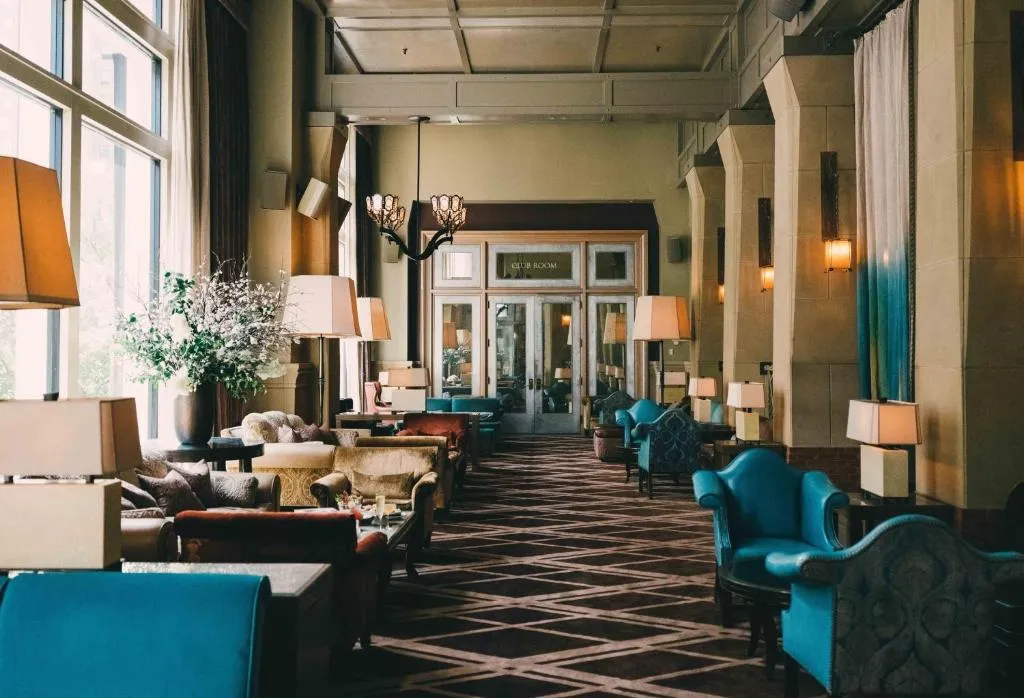 Elegant hotel lounge with blue and brown armchairs, multiple lamps, large windows, and patterned carpet.